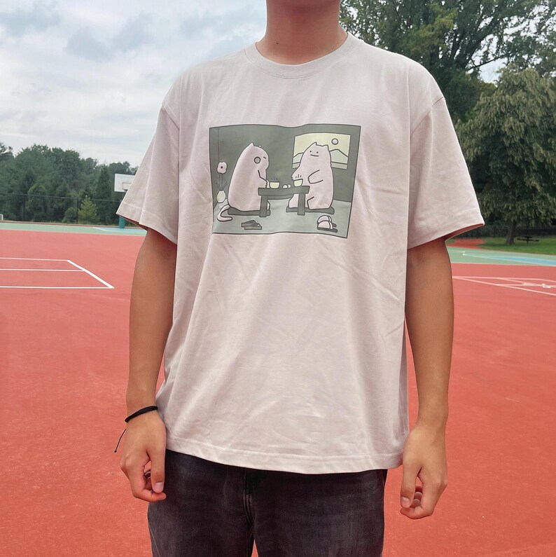 Puede incluir: Una camiseta de manga corta de color beige claro con un estampado gr&aacute;fico rectangular en el pecho. La ilustraci&oacute;n muestra a dos gatos de dibujos animados blancos sentados a una mesa bebiendo de tazas en una habitaci&oacute;n con una ventana.