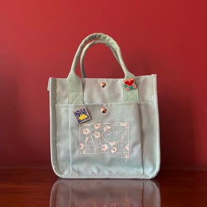 May include: A light green canvas tote bag with two white embroidered designs. One design is a cloud with the word "Greyo" inside. The other design is a landscape with two trees and flowers. The bag has two top handles and a small red flower pin on the front.