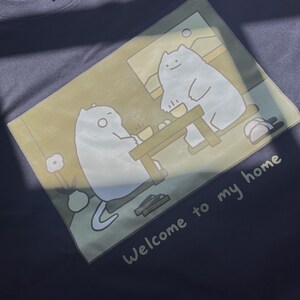 Puede incluir: Una camiseta gris oscuro con un estampado gr&aacute;fico de dos gatos blancos sentados a una mesa de madera en una habitaci&oacute;n. La ilustraci&oacute;n incluye una flor en un jarr&oacute;n y el texto "Welcome to my home" impreso en blanco bajo la escena.