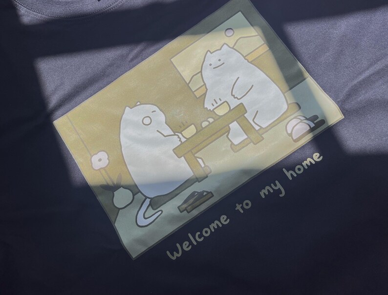 Puede incluir: Una camiseta gris oscuro con un estampado gr&aacute;fico de dos gatos blancos sentados a una mesa de madera en una habitaci&oacute;n. La ilustraci&oacute;n incluye una flor en un jarr&oacute;n y el texto "Welcome to my home" impreso en blanco bajo la escena.