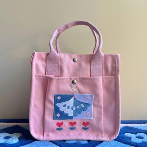 Puede incluir: Una bolsa de lona rosa con un dise&ntilde;o bordado azul y morado de un p&aacute;jaro con flores. La bolsa tiene dos bolsillos en la parte delantera y dos asas.