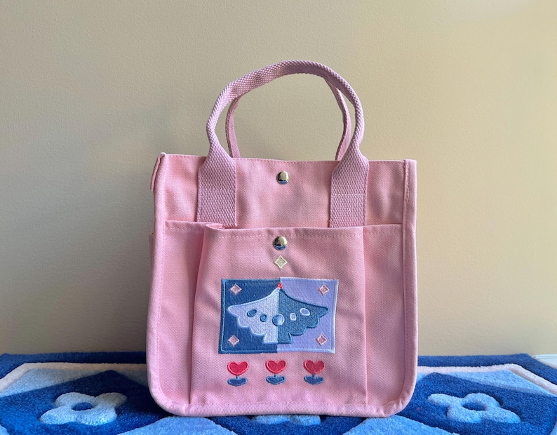 Puede incluir: Una bolsa de lona rosa con un dise&ntilde;o bordado azul y morado de un p&aacute;jaro con flores. La bolsa tiene dos bolsillos en la parte delantera y dos asas.