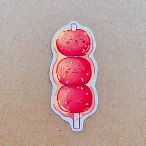 May include: A sticker featuring three red, round, smiling food items on a stick. Each item has white sesame seeds and is surrounded by a clear, bubbly outline. The sticker has a white border and is on a tan background.