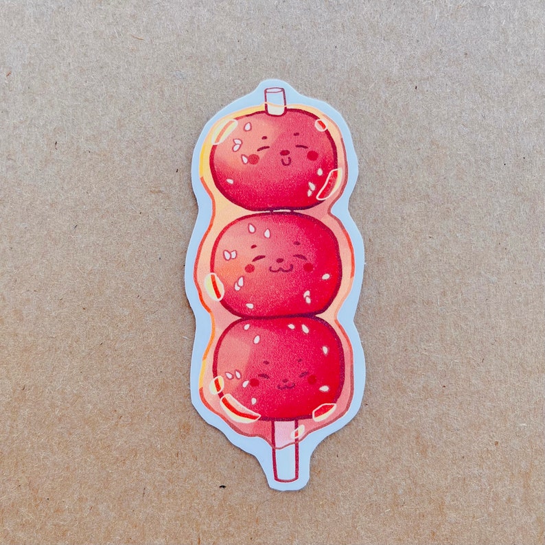 May include: A sticker featuring three red, round, smiling food items on a stick. Each item has white sesame seeds and is surrounded by a clear, bubbly outline. The sticker has a white border and is on a tan background.