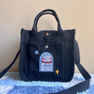 Puede incluir: Bolso tote de lona negro con un gato bordado en la parte delantera. El gato está mirando por una ventana con una luna y una estrella en el fondo.