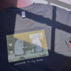 Puede incluir: Una camiseta de manga corta de color gris carb&oacute;n con un estampado gr&aacute;fico de dos gatos blancos de dibujos animados sentados a una mesa, con el texto "Welcome to my home" impreso debajo de la ilustraci&oacute;n. Tiene cuello redondo.