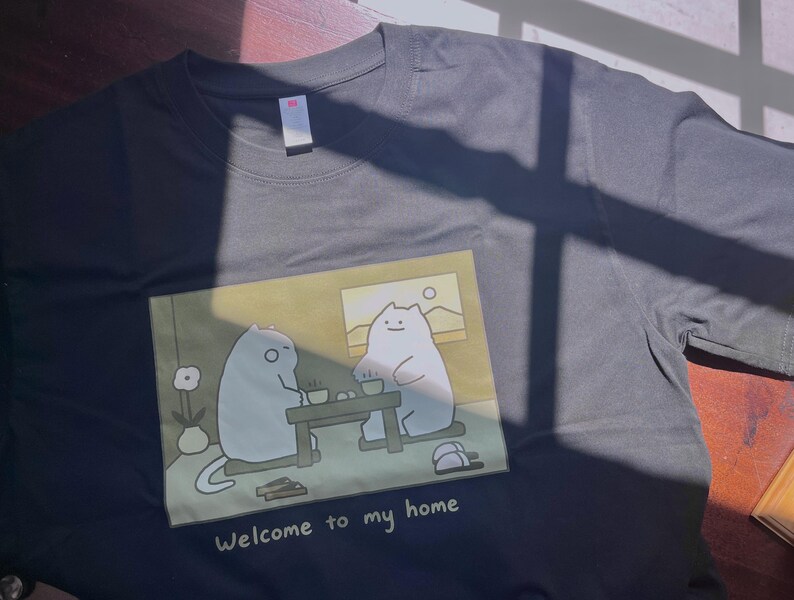 Puede incluir: Una camiseta de manga corta de color gris carb&oacute;n con un estampado gr&aacute;fico de dos gatos blancos de dibujos animados sentados a una mesa, con el texto "Welcome to my home" impreso debajo de la ilustraci&oacute;n. Tiene cuello redondo.