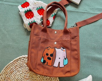 Puede incluir: Una bolsa de lona marrón con dos animales de dibujos animados que sostienen bolsas azules. La bolsa tiene una correa larga marrón y un asa marrón más corta.