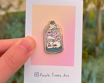 May include: Gold enamel pin featuring a glass bottle with a pink and purple gradient, clouds, a crescent moon, stars, and two small pink and white characters inside. The pin is on a white card with the text "Purple. Times. Art" in black.