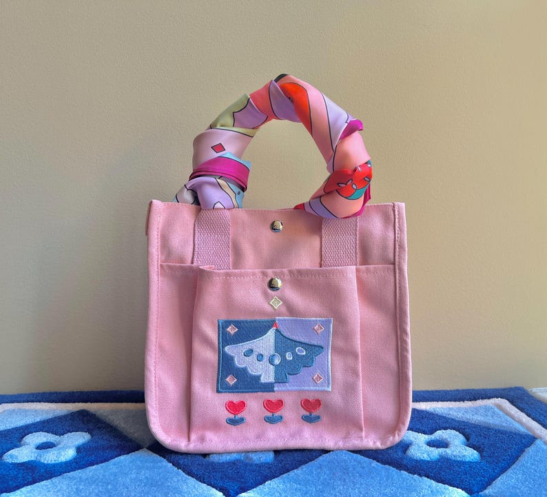 Puede incluir: Una bolsa de lona rosa con un asa de pa&ntilde;uelo de estampado colorido. La bolsa tiene un gran dise&ntilde;o bordado de un p&aacute;jaro azul y morado con tres corazones rojos debajo.