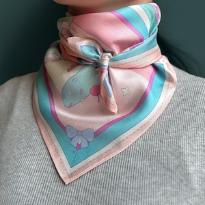 Peut inclure: Un foulard en soie à motifs roses et bleus avec un nœud noué au centre. Le foulard a un design fantaisiste avec un objet vert et blanc et une cerise rose.