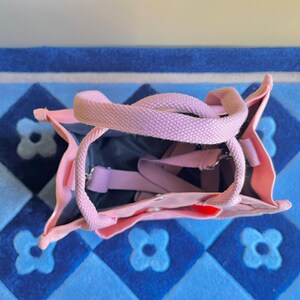 Puede incluir: Una bolsa de lona rosa con una correa de lona rosa claro. La bolsa est&aacute; abierta y el interior es visible. La bolsa est&aacute; sobre una alfombra de patr&oacute;n azul y blanco.