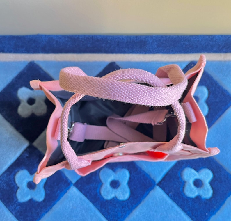 Puede incluir: Una bolsa de lona rosa con una correa de lona rosa claro. La bolsa est&aacute; abierta y el interior es visible. La bolsa est&aacute; sobre una alfombra de patr&oacute;n azul y blanco.