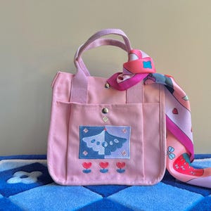 Puede incluir: Una bolsa de lona rosa con un asa de color morado claro y un dise&ntilde;o bordado en rosa y azul. La bolsa tiene un peque&ntilde;o bolsillo en la parte delantera y est&aacute; decorada con una bufanda estampada en rosa y blanco.