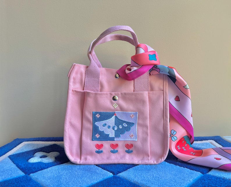 Puede incluir: Una bolsa de lona rosa con un asa de color morado claro y un dise&ntilde;o bordado en rosa y azul. La bolsa tiene un peque&ntilde;o bolsillo en la parte delantera y est&aacute; decorada con una bufanda estampada en rosa y blanco.