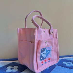 Puede incluir: Una bolsa de lona rosa con un dise&ntilde;o bordado azul y morado y tres corazones rojos. La bolsa tiene un asa superior y dos asas m&aacute;s cortas.
