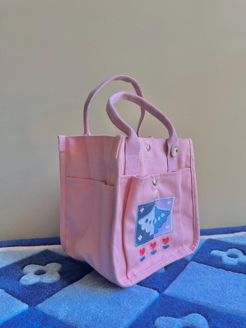 Puede incluir: Una bolsa de lona rosa con un dise&ntilde;o bordado azul y morado y tres corazones rojos. La bolsa tiene un asa superior y dos asas m&aacute;s cortas.