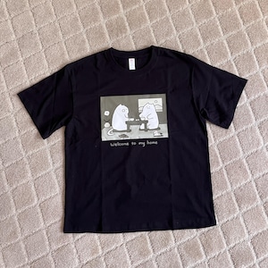 Puede incluir: Una camiseta negra de manga corta con un estampado gr&aacute;fico rectangular de dos gatos blancos de dibujos animados sentados a una mesa, con el texto "Welcome to my home" impreso debajo de la ilustraci&oacute;n en una fuente blanca sencilla.
