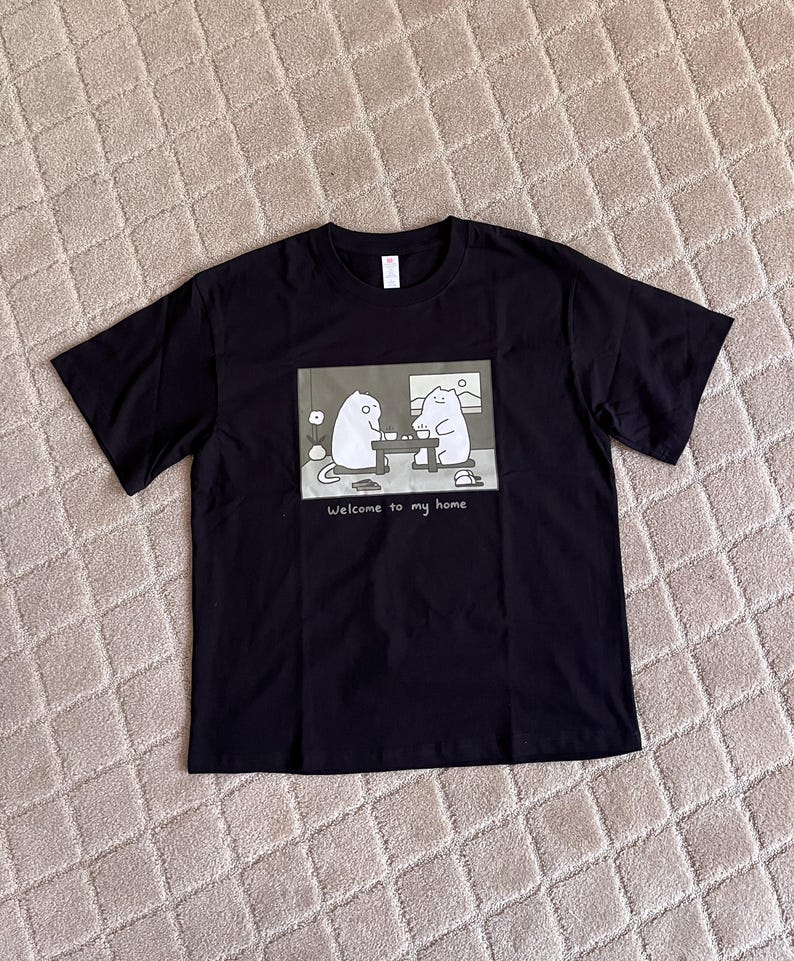 Puede incluir: Una camiseta negra de manga corta con un estampado gr&aacute;fico rectangular de dos gatos blancos de dibujos animados sentados a una mesa, con el texto "Welcome to my home" impreso debajo de la ilustraci&oacute;n en una fuente blanca sencilla.