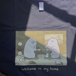 Puede incluir: Una camiseta negra con un estampado gr&aacute;fico rectangular de dos gatos blancos de dibujos animados sentados a una mesa baja con tazas de t&eacute;. El texto "Welcome to my home" est&aacute; impreso en blanco debajo de la ilustraci&oacute;n.