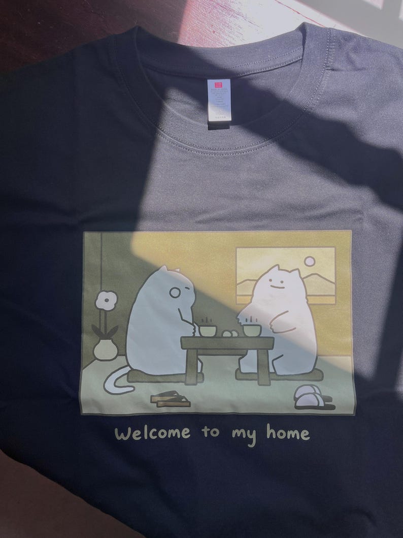 Puede incluir: Una camiseta negra con un estampado gr&aacute;fico rectangular de dos gatos blancos de dibujos animados sentados a una mesa baja con tazas de t&eacute;. El texto "Welcome to my home" est&aacute; impreso en blanco debajo de la ilustraci&oacute;n.