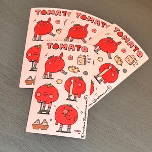 May include: A set of five pink and white stickers featuring a cartoon tomato character in different poses. The stickers have the word "TOMATO" printed in black at the top. The tomato character is wearing grey socks and shoes. The stickers are on a grey surface.