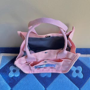 Puede incluir: Una bolsa de lona rosa con una ilustraci&oacute;n de dibujos animados de una casa con un techo azul y una chimenea blanca. La bolsa tiene dos bolsillos en la parte delantera y dos asas.
