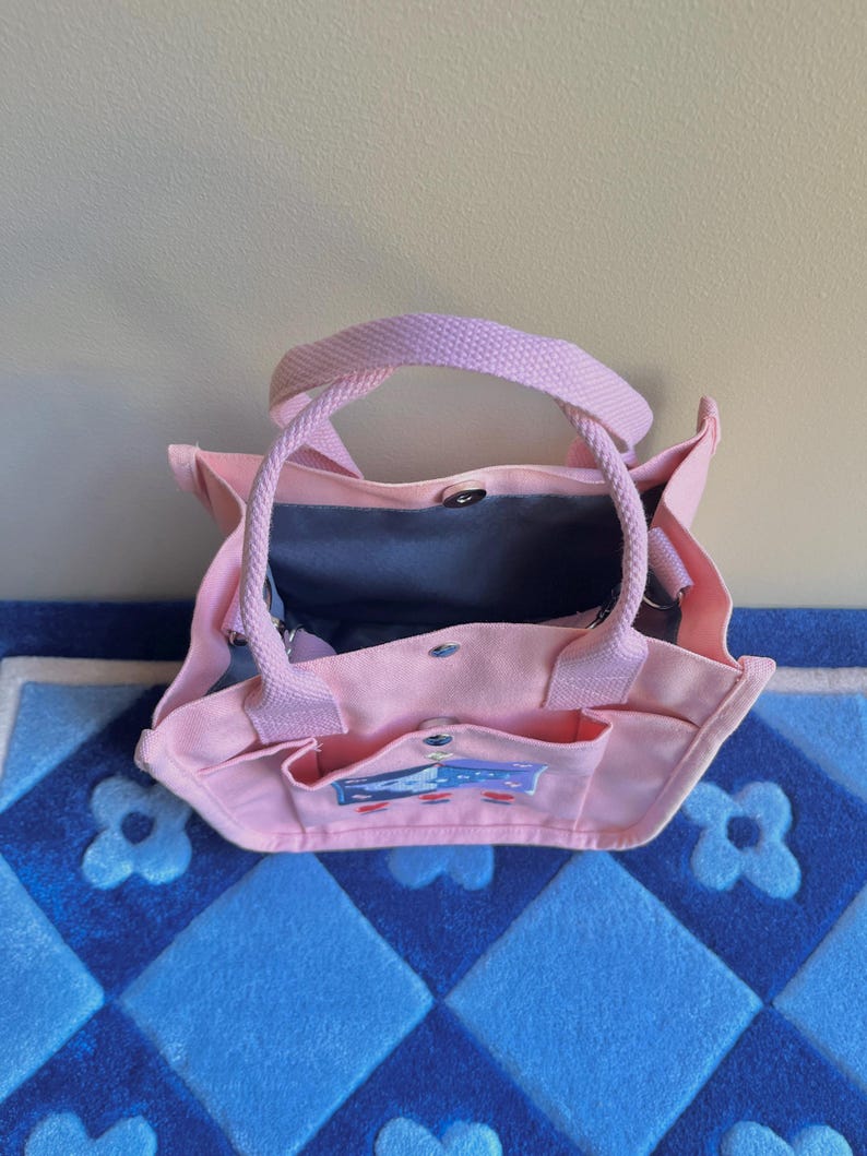 Puede incluir: Una bolsa de lona rosa con una ilustraci&oacute;n de dibujos animados de una casa con un techo azul y una chimenea blanca. La bolsa tiene dos bolsillos en la parte delantera y dos asas.