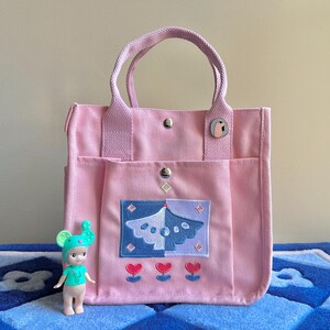 Puede incluir: Una bolsa de lona rosa con un dise&ntilde;o bordado en azul y blanco de un &aacute;rbol con corazones en la parte inferior. La bolsa tiene dos asas superiores y un bolsillo con cremallera en la parte delantera.