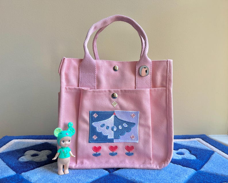 Puede incluir: Una bolsa de lona rosa con un dise&ntilde;o bordado en azul y blanco de un &aacute;rbol con corazones en la parte inferior. La bolsa tiene dos asas superiores y un bolsillo con cremallera en la parte delantera.