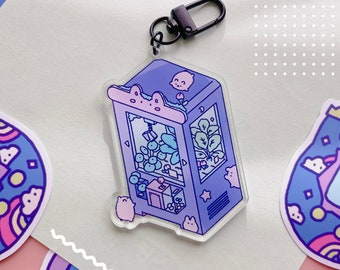 May include: A purple and pink claw machine keychain with a black metal ring. The claw machine is filled with small, round, blue and white objects. There are also small, green plants and a small, white animal inside the claw machine.