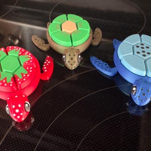 May include: Three colorful turtle toys with segmented shells. The turtle on the left has a red shell with green and white accents. The turtle in the center has a green shell with a brown center. The turtle on the right has a blue shell with a gray center.