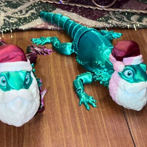 May include: Two teal green lizard figurines wearing Santa hats and white beards. The lizards are posed in a walking position.