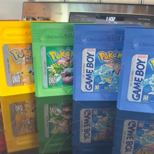 May include: A collection of colorful Game Boy cartridges featuring the Pokémon series. The cartridges are red, yellow, green, and blue, each with unique game art and the "Pokémon" and "Game Boy" logos.