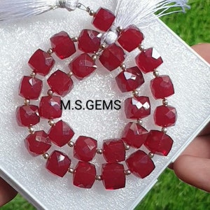 May include: A strand of red gemstone beads with a silver clasp and white tassel. The beads are faceted and have a square shape. The beads are displayed in a white box with the text "M.S. GEMS" printed on the box.