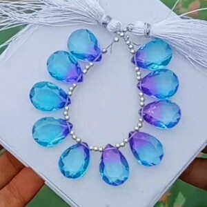 May include: A strand of blue and purple teardrop-shaped gemstone beads strung on a silver chain with white tassels at each end.