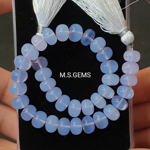 May include: A strand of pale blue and pink oval-shaped gemstone beads with a white tassel on each end. The beads are arranged in a circle on a black surface. The text "M.S.GEMS" is visible in the center of the image.