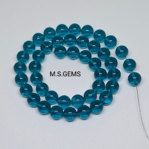 May include: A strand of teal blue glass beads. The beads are round and have a smooth, polished finish. M.S. GEMS is printed on the white background.