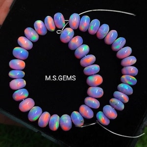 Può includere: Una collana di perle di opale iridescenti in tonalità rosa, viola e blu. Le perle sono infilate su un filo d'argento e disposte in un motivo circolare. Le perle hanno un diametro di circa 10 mm. M.S. GEMS è stampato sullo sfondo nero.