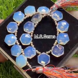 Op de afbeelding: Een streng iriserende opaal edelsteen kralen met een zilveren metalen ketting en een kleurrijke kwast. De kralen zijn gefacetteerd en hebben een zeshoekige vorm. De streng wordt getoond op een zwart oppervlak met de tekst "M.S. Gems".