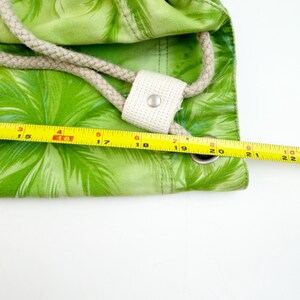 May include: A green drawstring bag with a palm leaf pattern. The bag has a beige rope drawstring and a white fabric reinforcement with a metal grommet. A yellow measuring tape shows the bag is approximately 22 inches wide.