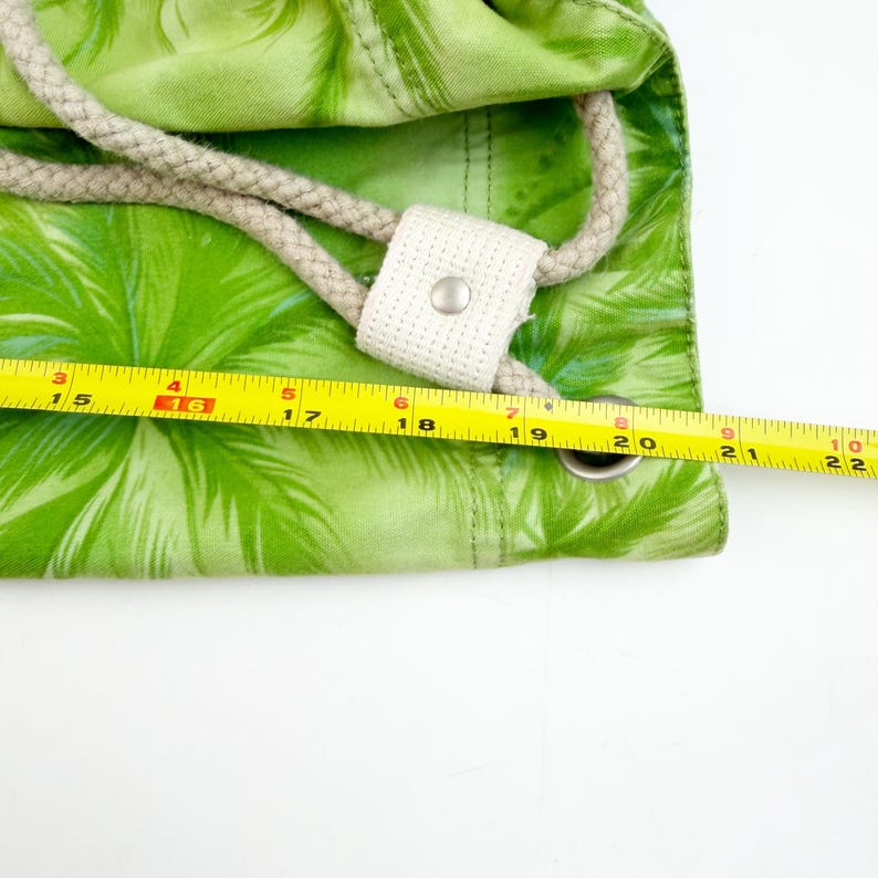 May include: A green drawstring bag with a palm leaf pattern. The bag has a beige rope drawstring and a white fabric reinforcement with a metal grommet. A yellow measuring tape shows the bag is approximately 22 inches wide.