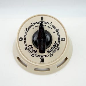 May include: A round, cream-colored kitchen timer with a black central knob. The face features black numerals from 0 to 55, tick marks, and the text "Clock Watcher" and "TURN PAST DOT". The base has three rectangular cutouts along the edge.
