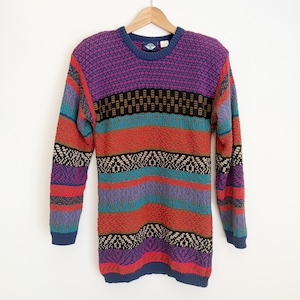 May include: A long-sleeve crew neck sweater featuring a multi-colored horizontal striped pattern in shades of purple, red, teal, black, and tan. The knit garment has a ribbed collar, cuffs, and hem, and is displayed on a wooden clothes hanger.