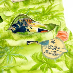 May include: Close-up of a lime green tote bag with a tropical palm tree pattern. The bag has a zippered pocket revealing a colorful interior. A tag reads "Fly to TBI Bora Bora."