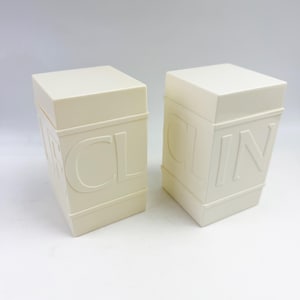 May include: Two off-white rectangular containers with lids, each featuring embossed lettering on the side. The left container displays the letters CL, while the right container displays the letters IN. Both items have a smooth, solid-colored finish.