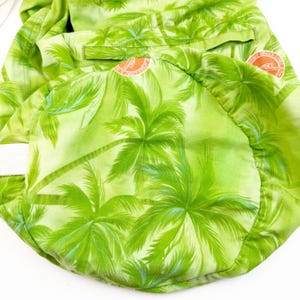 May include: Green shorts with a vibrant palm tree pattern. The shorts feature a white elastic waistband and two small orange circular patches. The fabric has a light green gradient, with darker green palm tree illustrations.