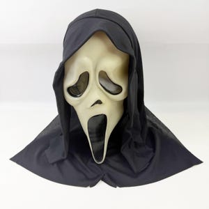 May include: A Scream mask, featuring a pale, elongated face with large, oval eye openings and a wide, open mouth. The mask is framed by a black hood, creating a stark contrast against the off-white face. The design is a classic Halloween costume.