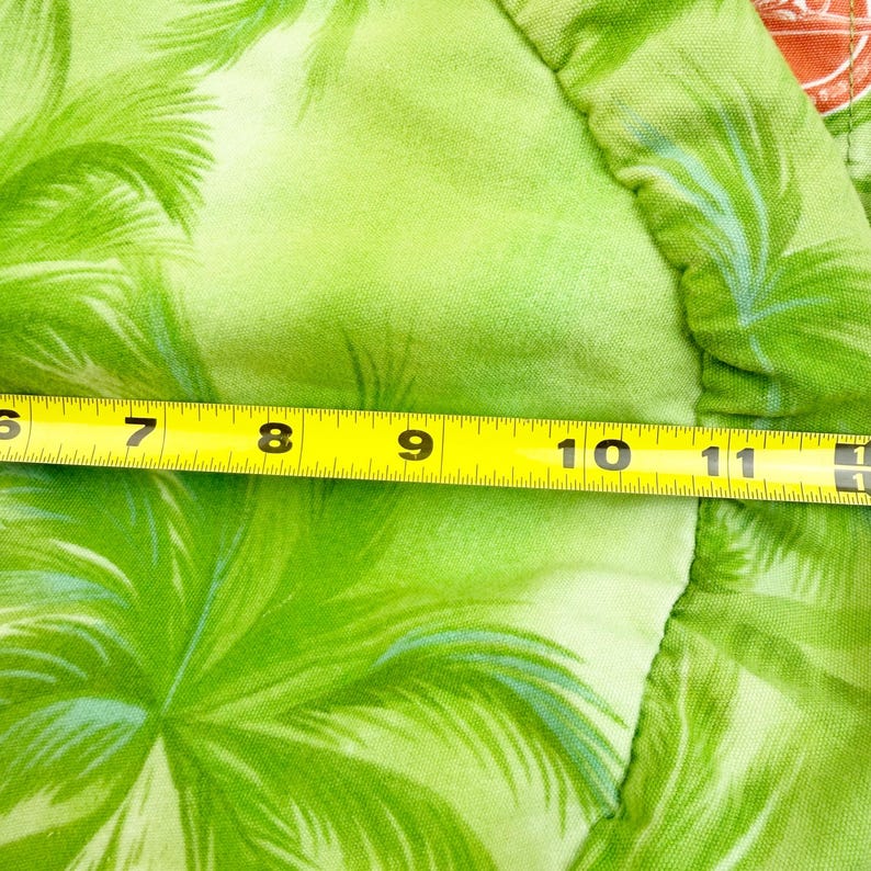 May include: Close-up of a bright green fabric with a tropical palm tree pattern. A yellow measuring tape shows a measurement of approximately 10 inches. The fabric has a light green background with darker green palm fronds.