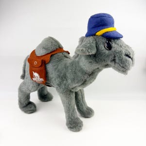 May include: A gray plush camel toy wearing a blue cap with a yellow band. The camel features a brown felt saddle with a small pocket and a white logo that reads Camel Korps. The stuffed animal has large black eyes and a soft, textured gray fur.